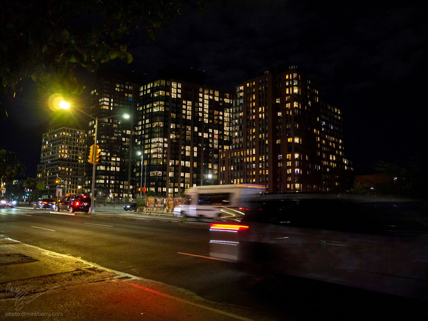 A very yellow hued photograph of a street at night, where moving cars are slightly blurred. Large blocky apartment buildings 